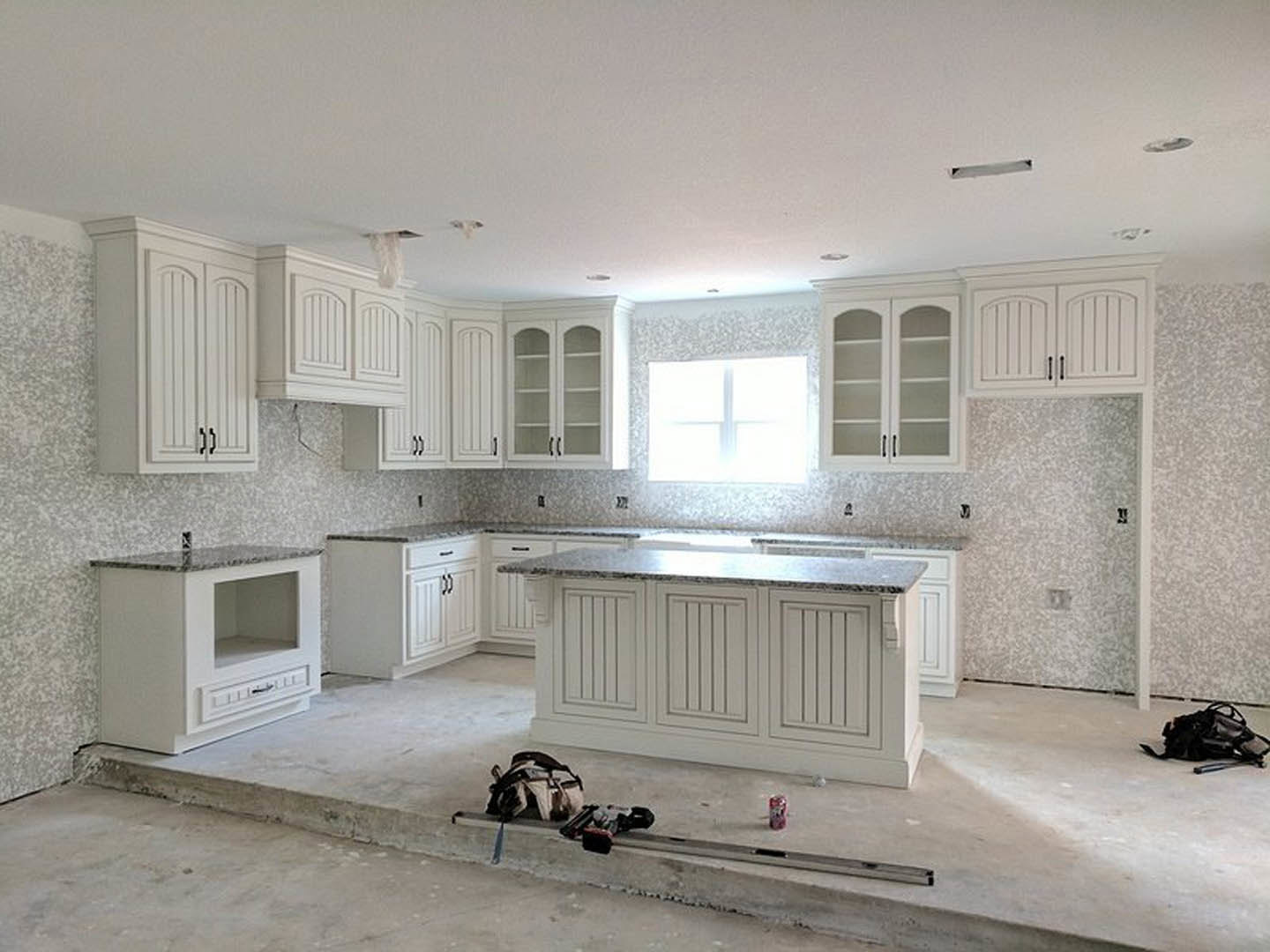 White kitchen with marble countertops, glass-front cabinets, tile backsplash, stainless steel sink, and a black bag on the floor near cabinetry.