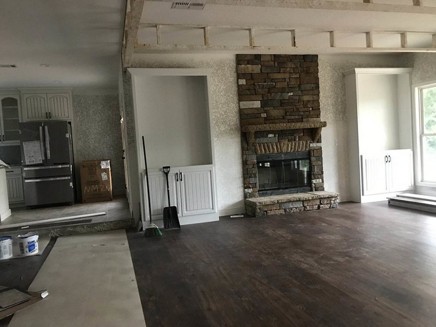 Living room featuring a stone fireplace, wood flooring, large window with glass panel, refrigerator with paper attached, broom and stick leaning against a white wall, cabinetry
