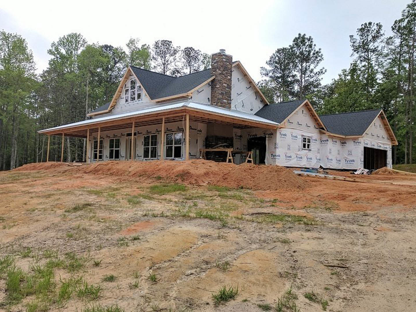 Partially built house with exposed stone pillar, white-framed window, and unfinished roof, set in a grassy dirt field with piles of earth; Karen Blixen Museum visible in the