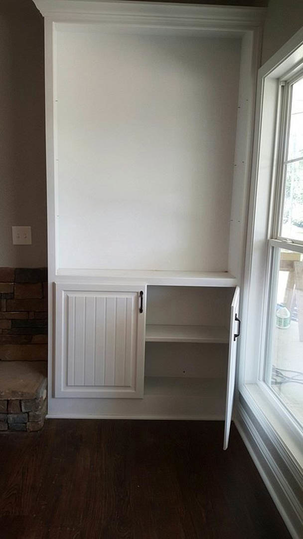 White shaker-style cabinet against a white wall, dark hardwood flooring with white baseboard trim, stone accent wall partially visible, open white door leading to adjacent room.