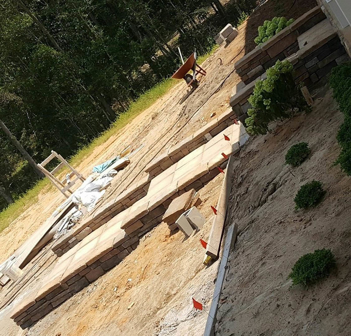 Partially built home foundation surrounded by grass, wheelbarrow, wooden chair, and green bushes; construction materials scattered on site.