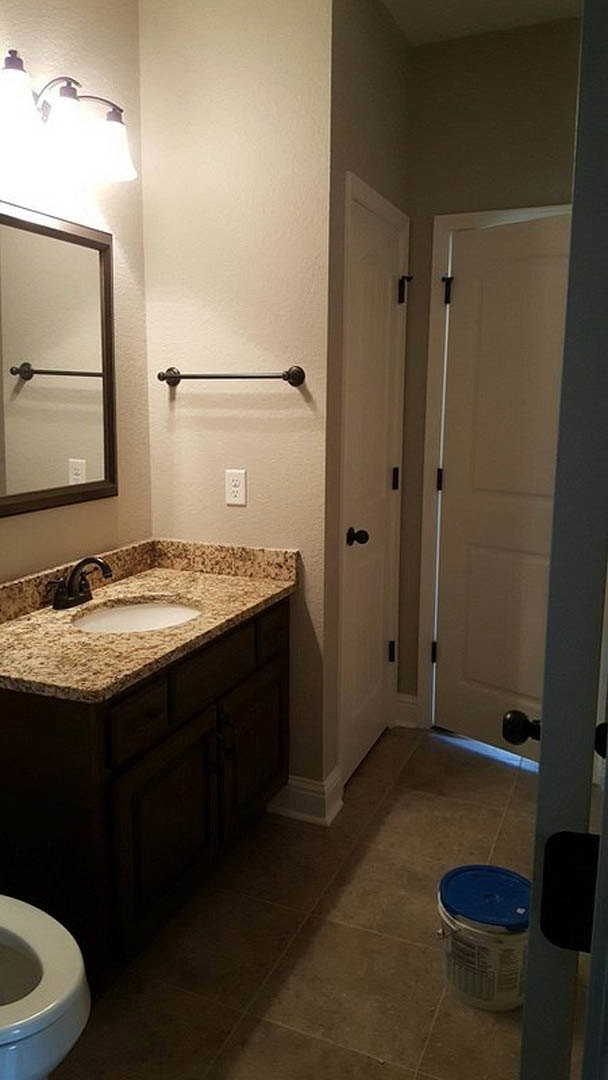 Modern bathroom featuring a white ceramic sink with chrome faucet, rectangular wall-mounted mirror, light gray tile walls, and partial view of a toilet and door frame