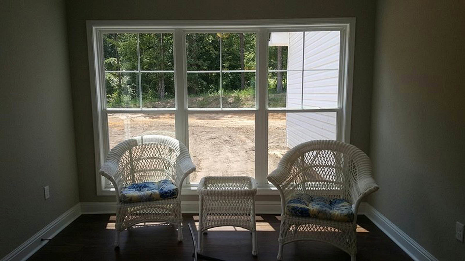 Two wicker chairs with cushions positioned in a bright room, natural light streaming through a large window with blinds, hardwood flooring and neutral walls visible.