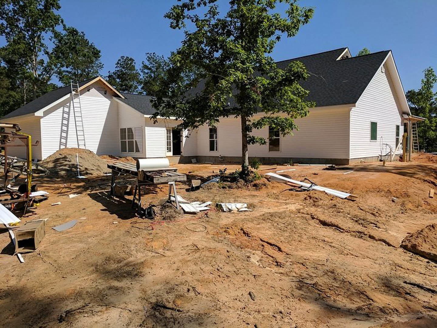 Framed house under construction with exposed wood, sandy soil, white pipe, and mature tree in front yard