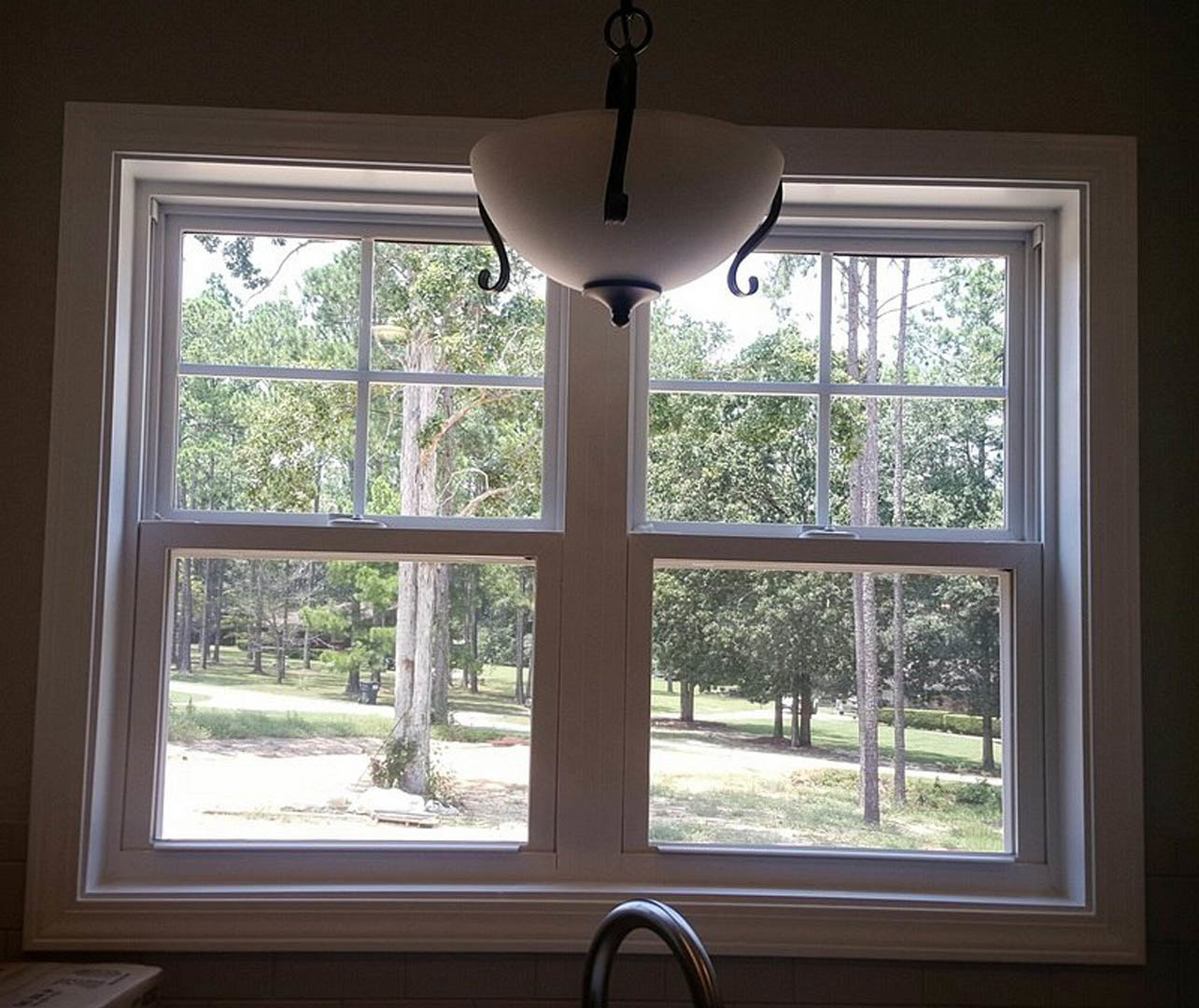 Kitchen interior featuring a large window with views of trees and park, pendant light fixture hanging from ceiling, neutral wall finishes, and window blinds.
