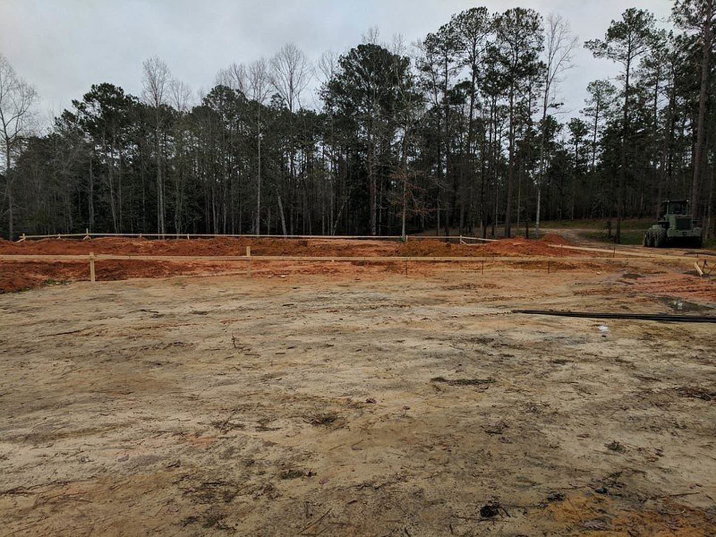 Dirt lot bordered by a wooden fence, scattered trees in the background, tractor with large wheel parked on soil, black object resting near rail in foreground, cloudy sky overhead