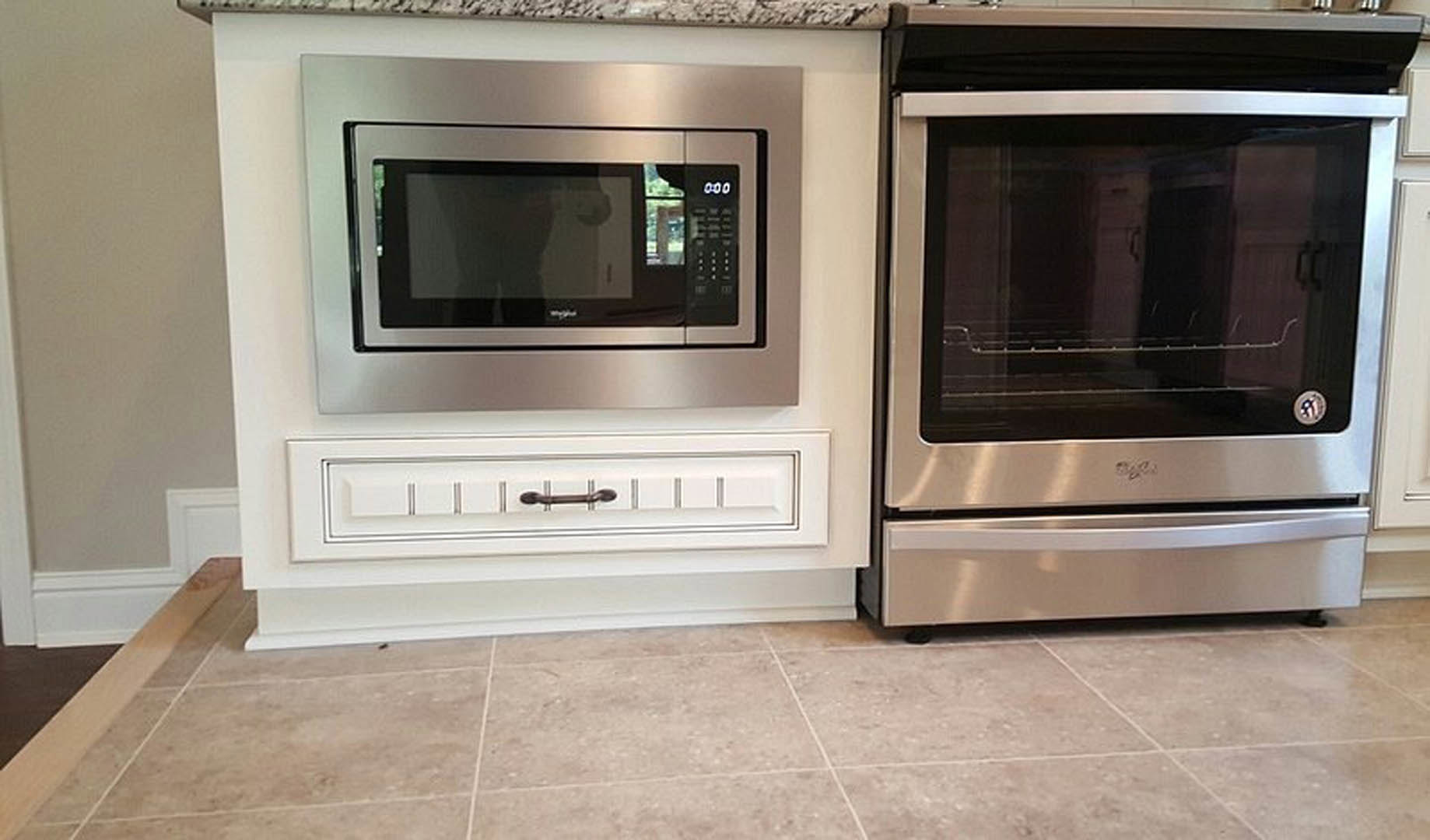 Stainless steel microwave and oven built into white cabinetry, tile flooring, brushed metal handles, and black accent lines