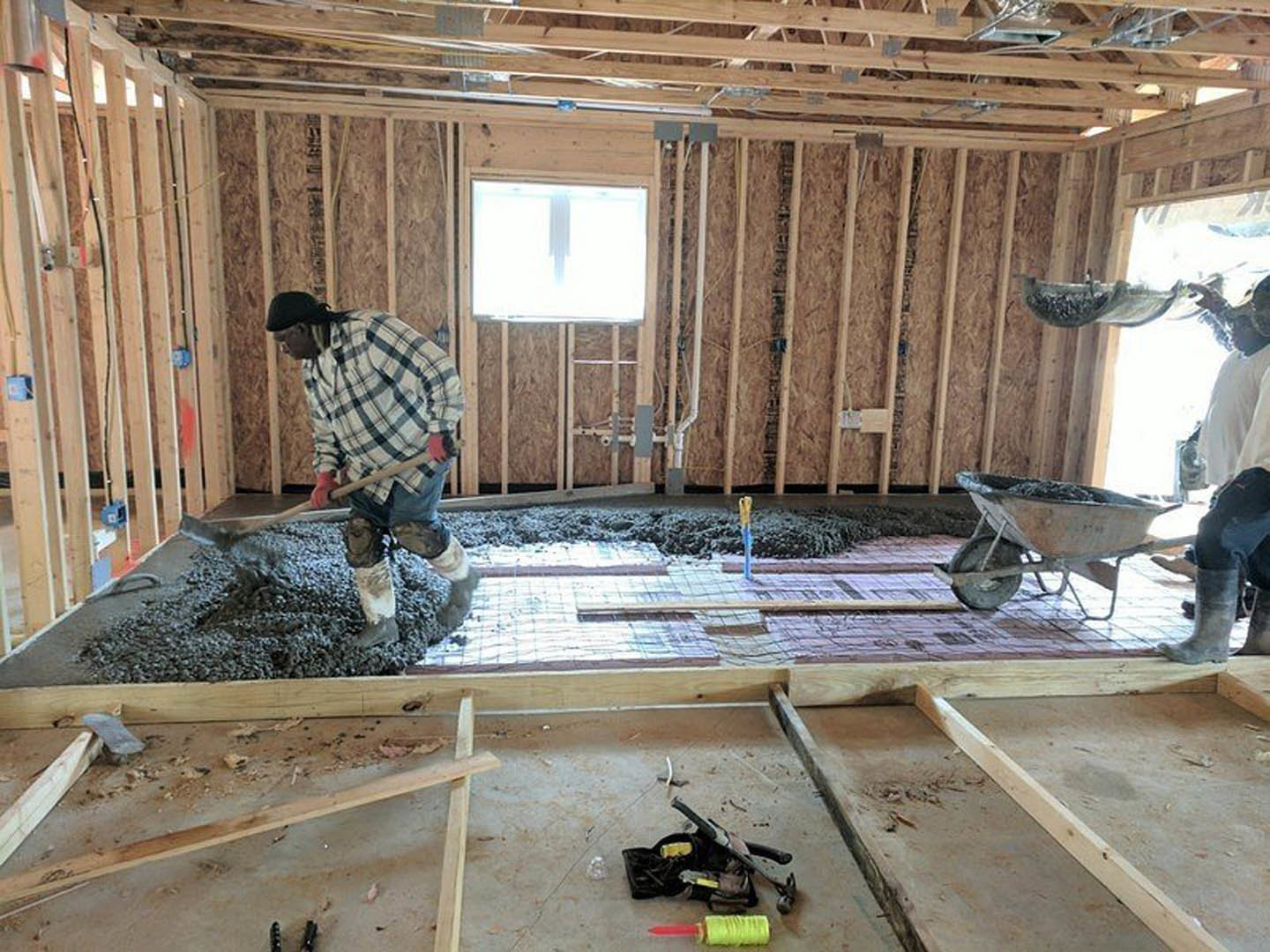 Man in blue shirt shoveling cement on unfinished floor, surrounded by lumber, construction tools, wheelbarrow, and building insulation inside residential home under remodel.