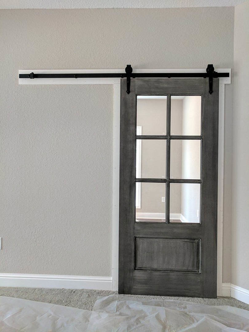 Black sliding barn door with glass panes, white cloth draped on light wood floor, adjacent plaster wall and window with metal handle.