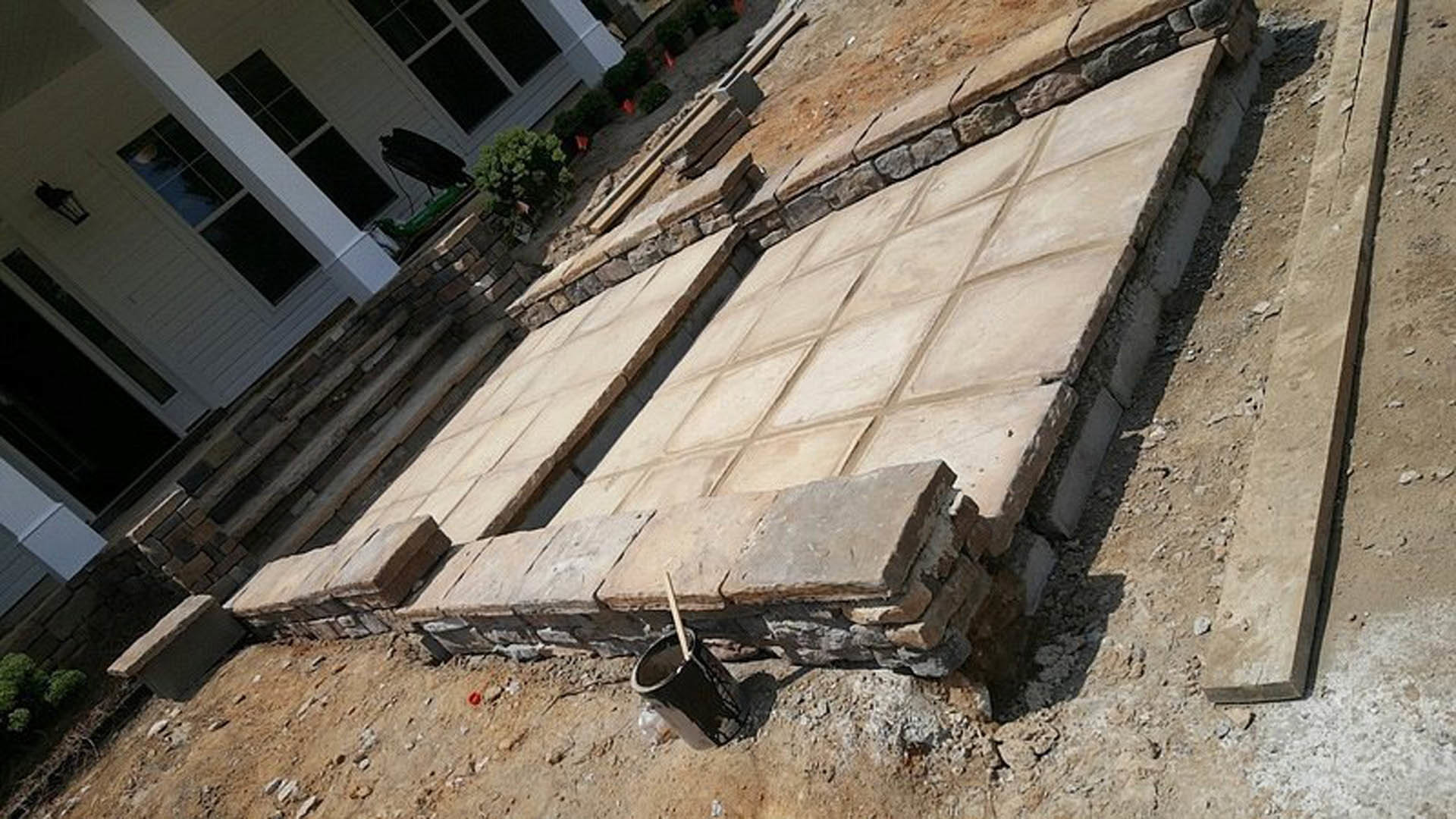 Stone block patio under construction with tiered steps, paint bucket and wooden stick nearby, leafy tree beside white house featuring porch and window