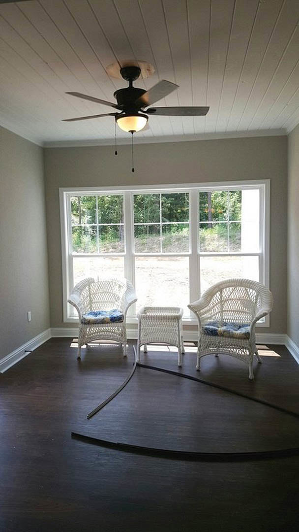 Wicker chairs with white cushions arranged around a wooden table in a bright room featuring large windows, ceiling fan, and light-colored flooring.