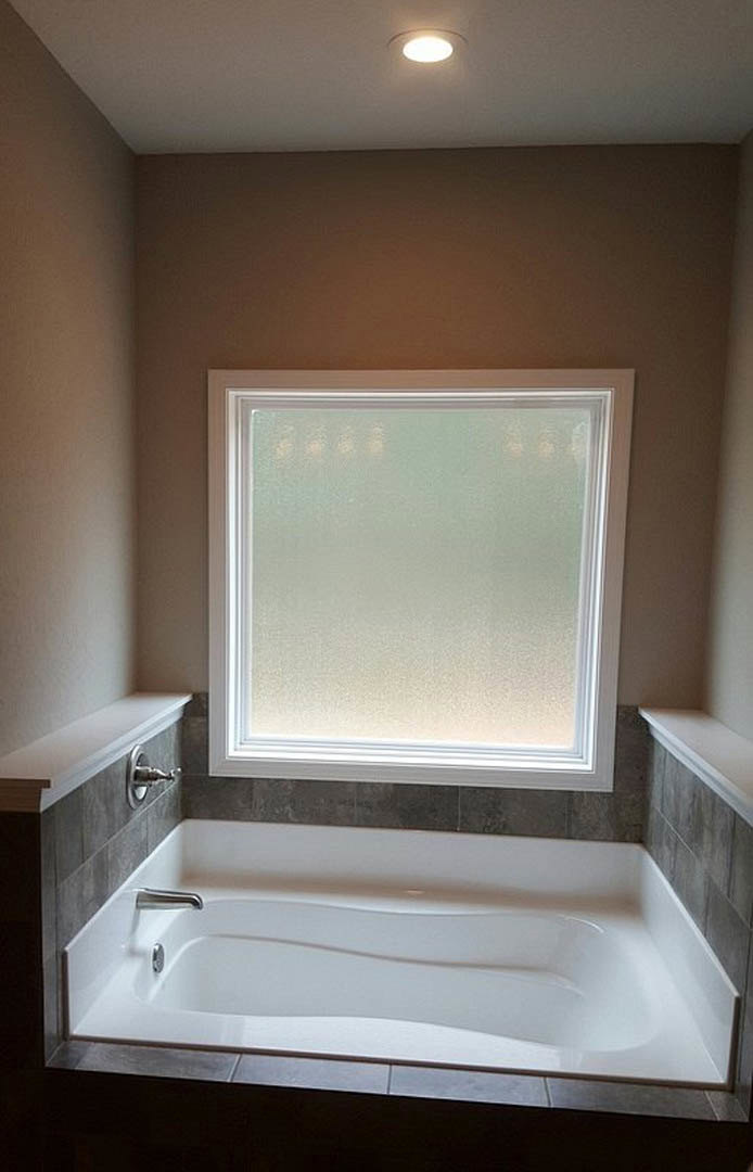 White freestanding bathtub beneath a large window in a bright bathroom with light walls and chrome fixtures
