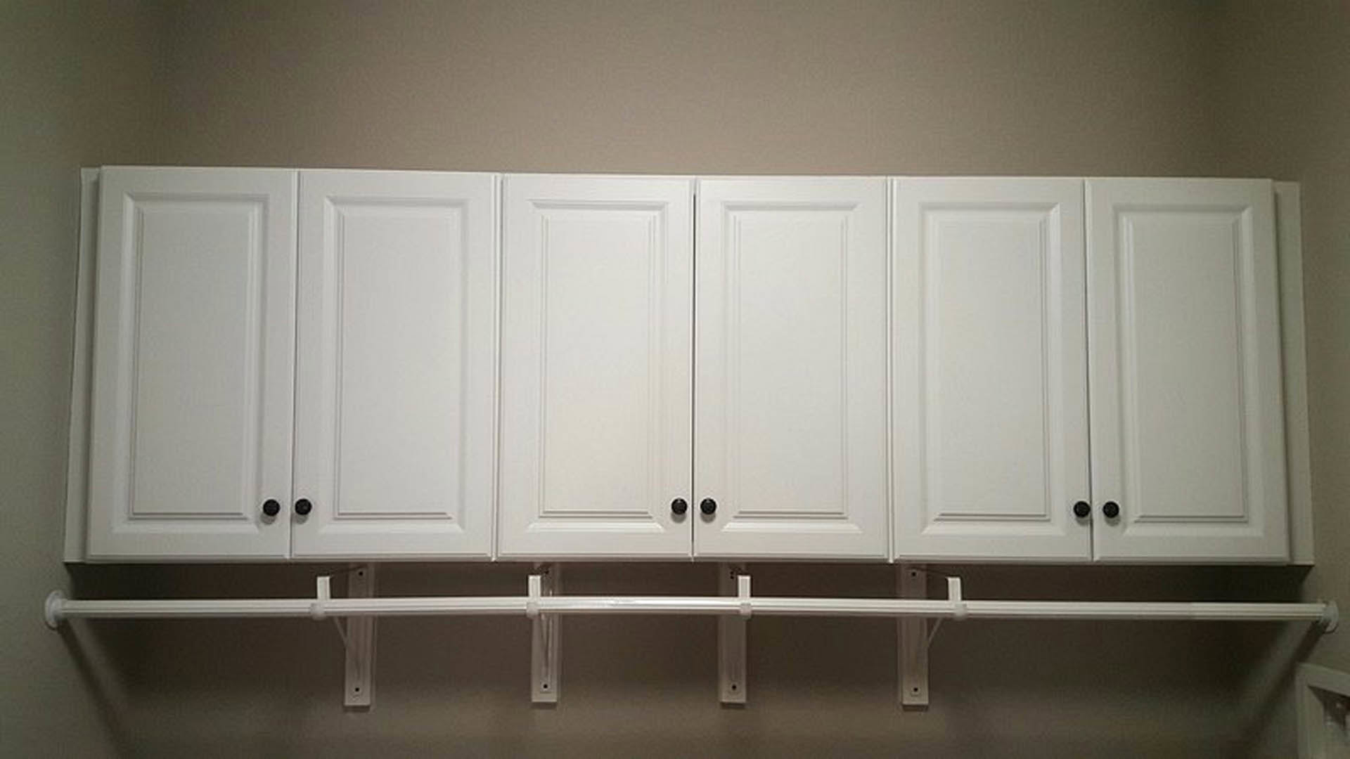 White cabinetry with black knobs mounted above a white shelf and rod against a light-colored wall.