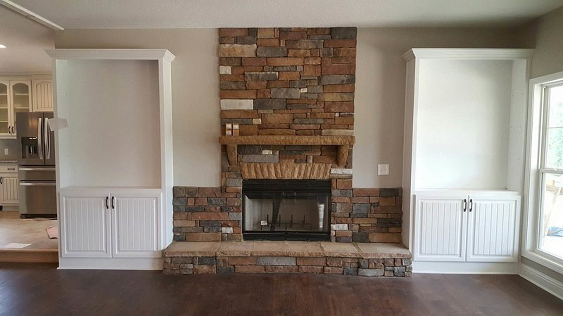 Modern den with brick fireplace, hardwood floors, white cabinetry with black handles, and large glass window; person standing near window, wood surface and refrigerator visible.