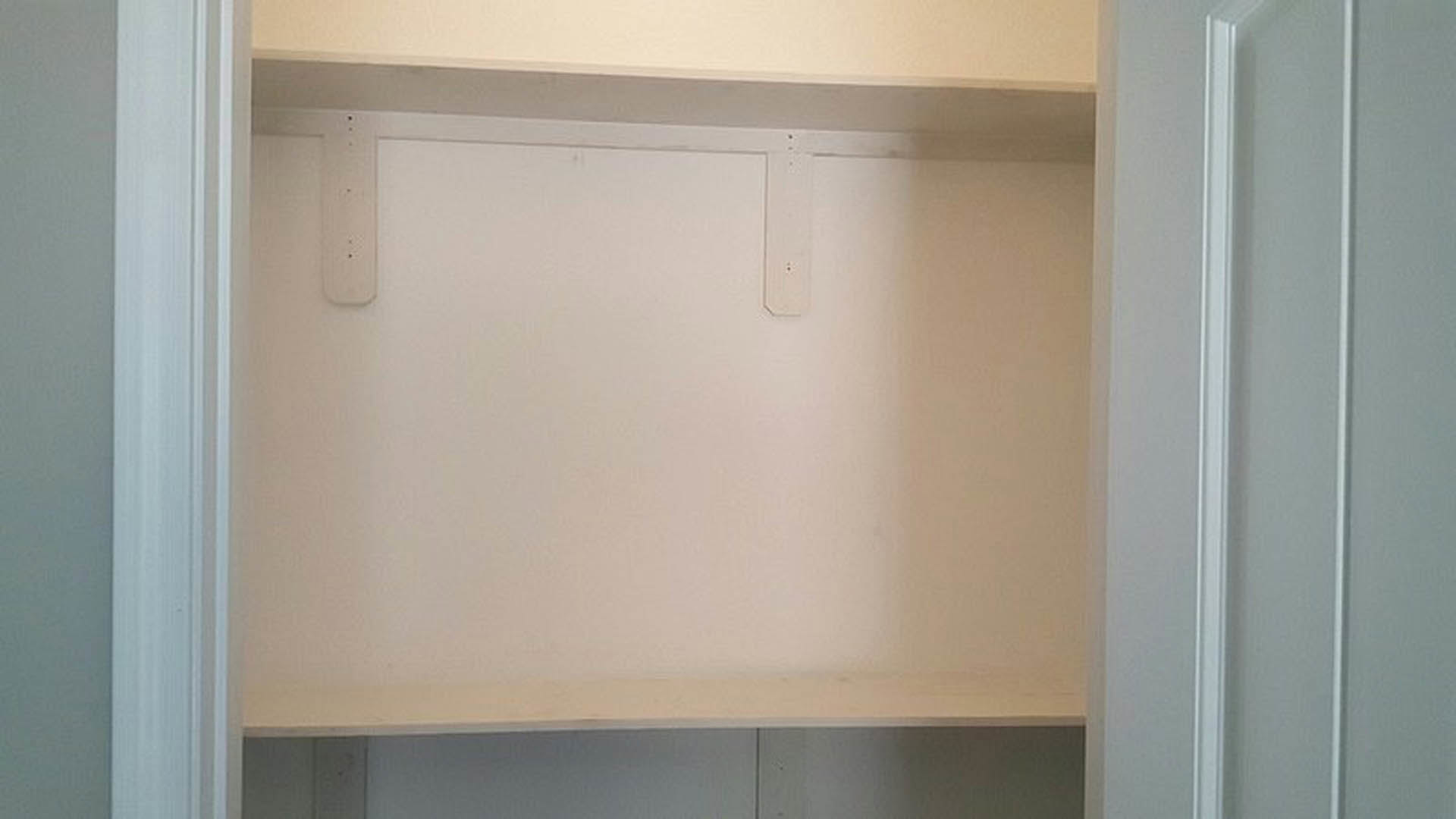 White closet shelf against a white plaster wall, black and white frame above, white door with small hole, blue background visible.