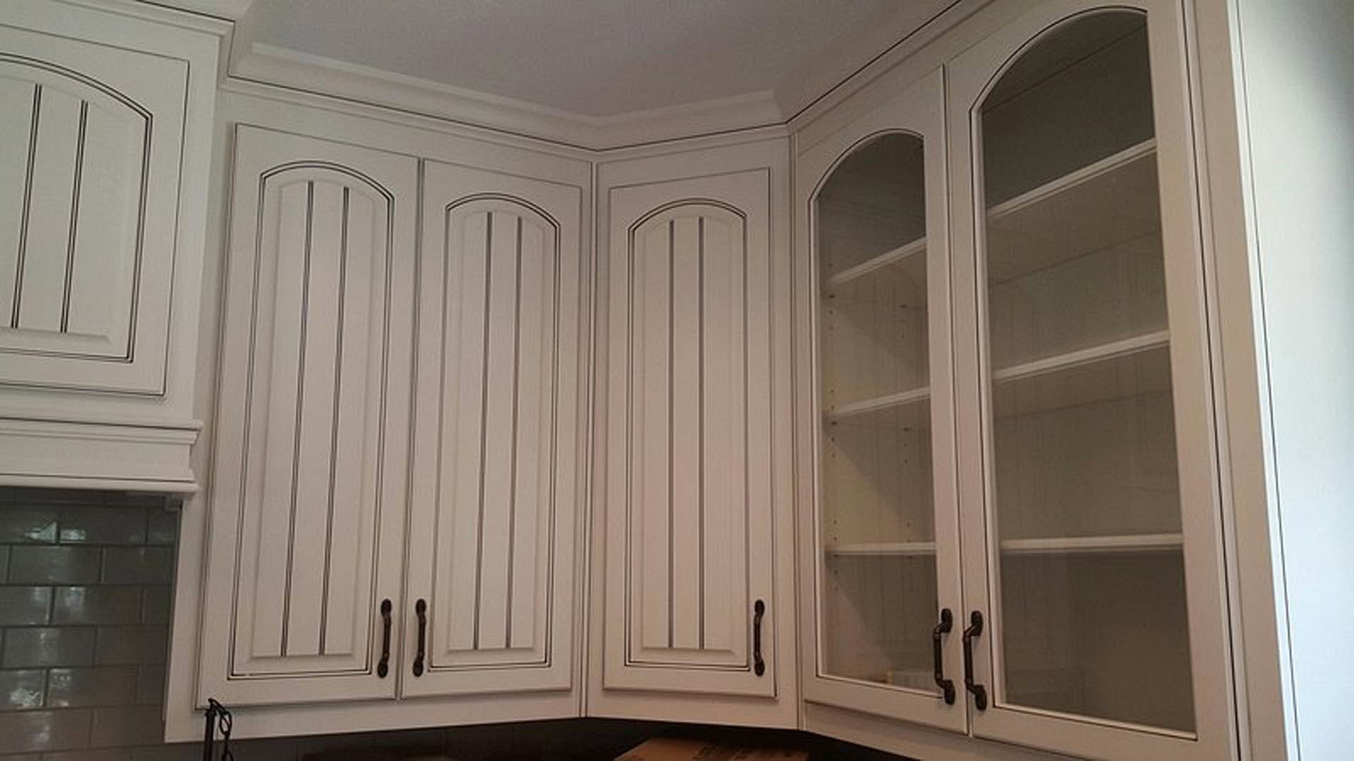 White kitchen cabinets with glass doors, brushed metal handles, white tile backsplash, crown molding, and wood flooring