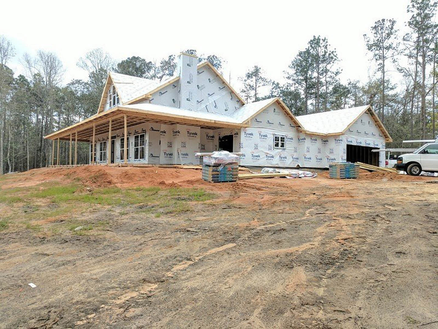 Partially built house with white insulation wrap, expansive covered porch, exposed roof framing, white van parked nearby, dirt yard with stacked boxes, tree and blue sky in