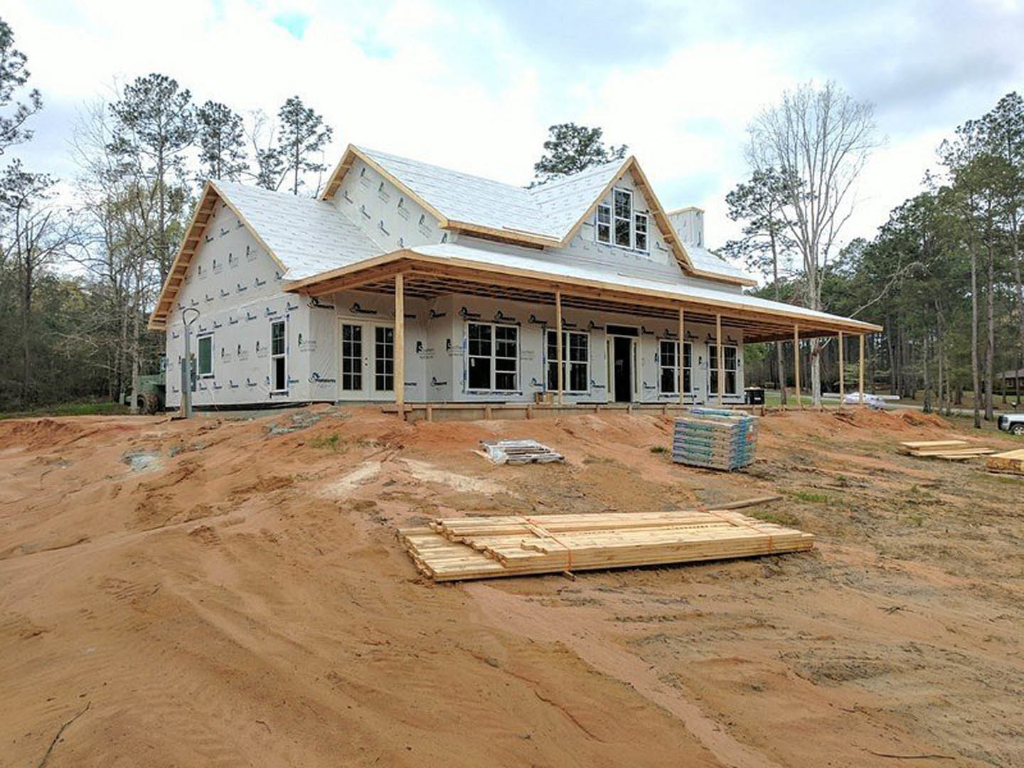 Partially built house with exposed wooden framing and roof, surrounded by dirt and construction materials, Karen Blixen Museum visible in background under cloudy sky.
