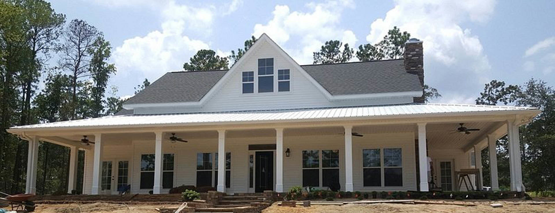 White siding house with wide covered porch, white columns, multiple windows with white frames, surrounded by trees and lawn under partly cloudy sky