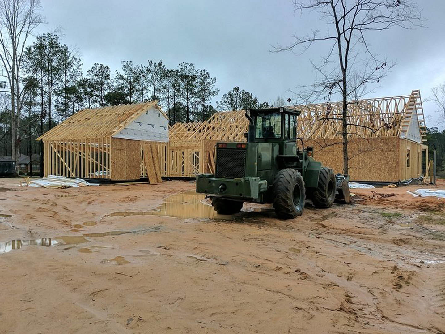 Green tractor with large tires parked on muddy ground beside a wooden structure with a sloped roof, tree visible next to the building under an open sky.