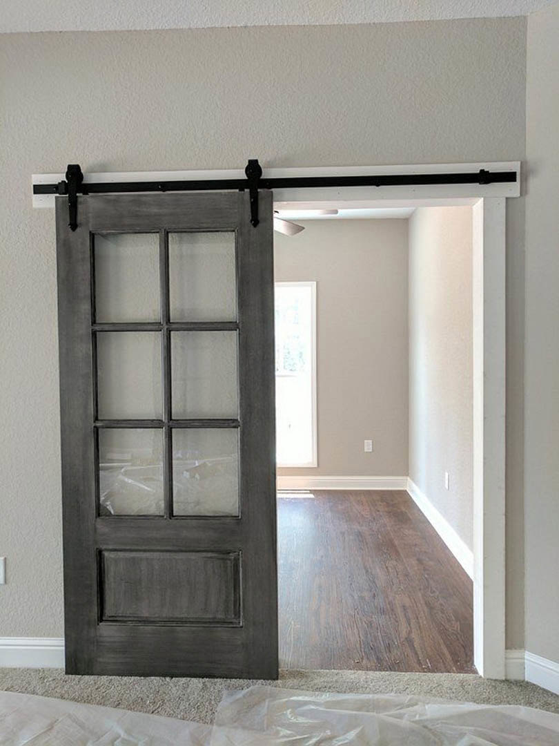 Black sliding barn door with matte hardware on dark wood flooring, white walls, and a whiteboard with handwritten notes; white plastic sheet partially covers carpet near window.