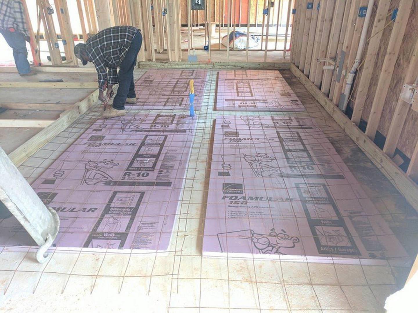 Man installing wooden flooring in custom home, holding a plank near white tubing, blueprint spread on subfloor, unfinished interior with construction materials visible.