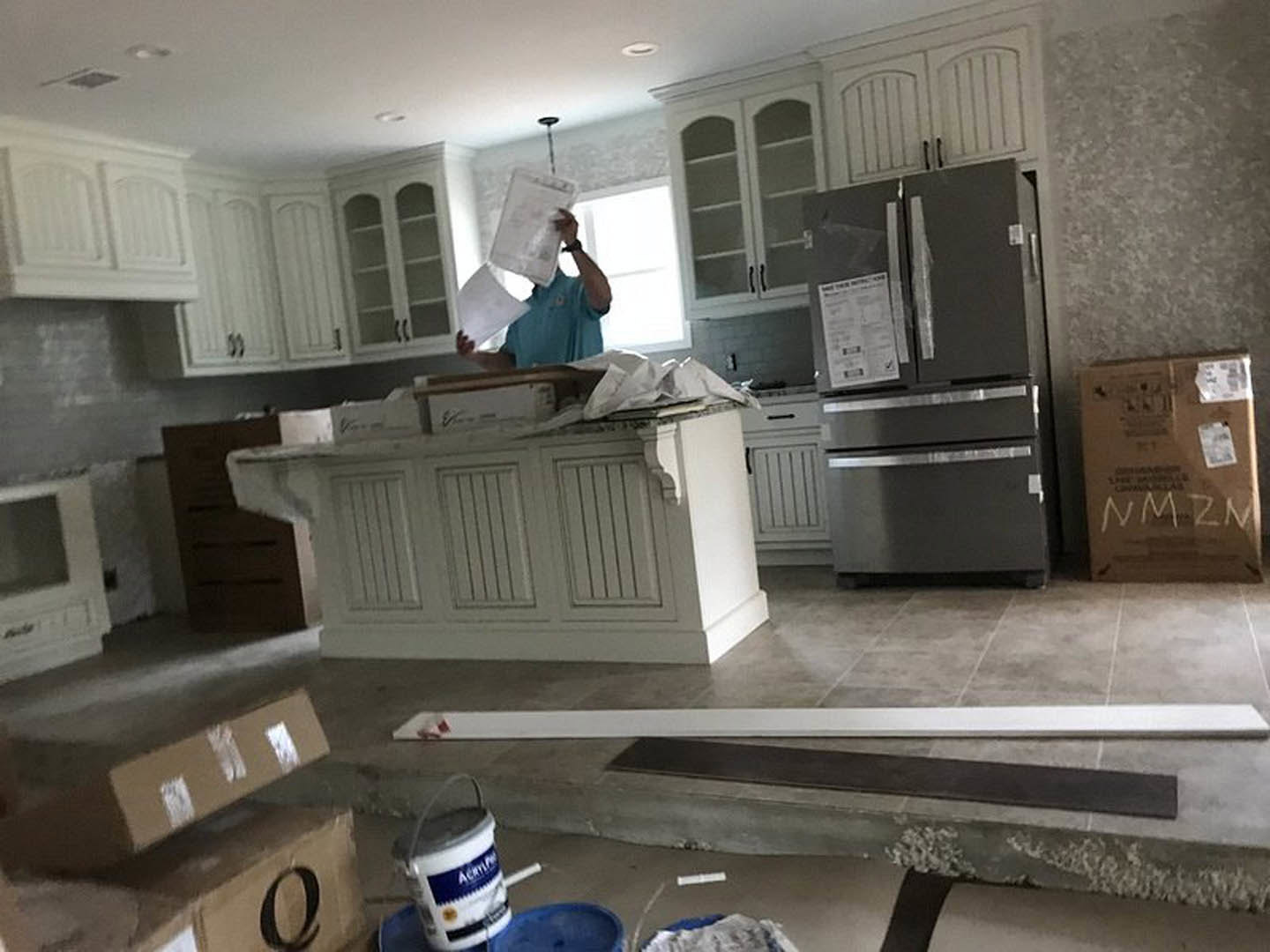 Man standing in kitchen with white cabinets, stainless refrigerator displaying a paper, brown box with white lettering on countertop, white bucket with handle, and papers held in