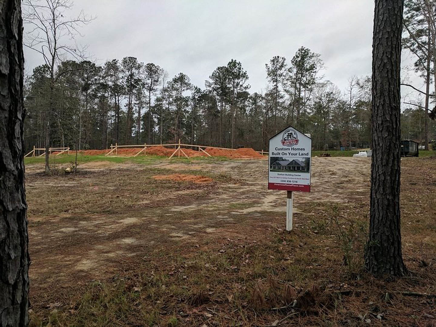 Wooden post with rectangular sign displaying house graphic, set in grassy clearing bordered by tall trees and dense forest under cloudy sky