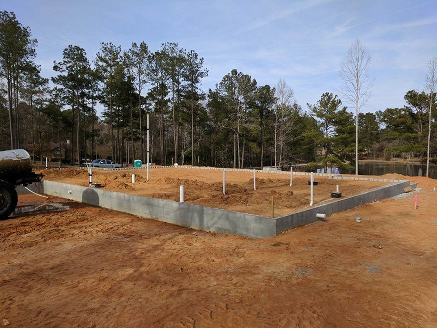 Concrete house foundation with white support poles, exposed soil, leafless tree, and wooded area in background under cloudy sky