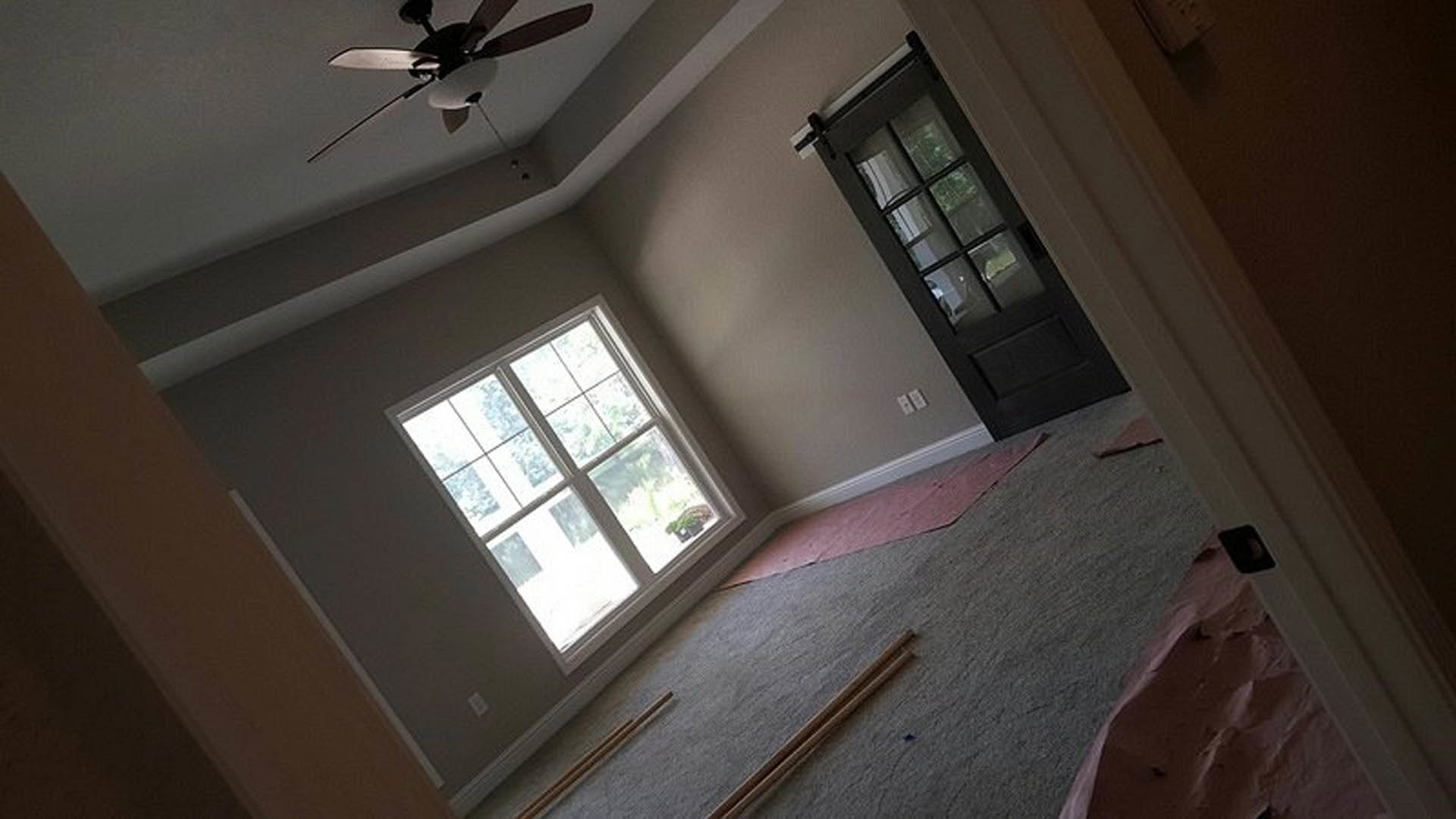 Bedroom with light wood flooring, white plaster walls, ceiling fan with integrated light, large window, and glass-paneled sliding door