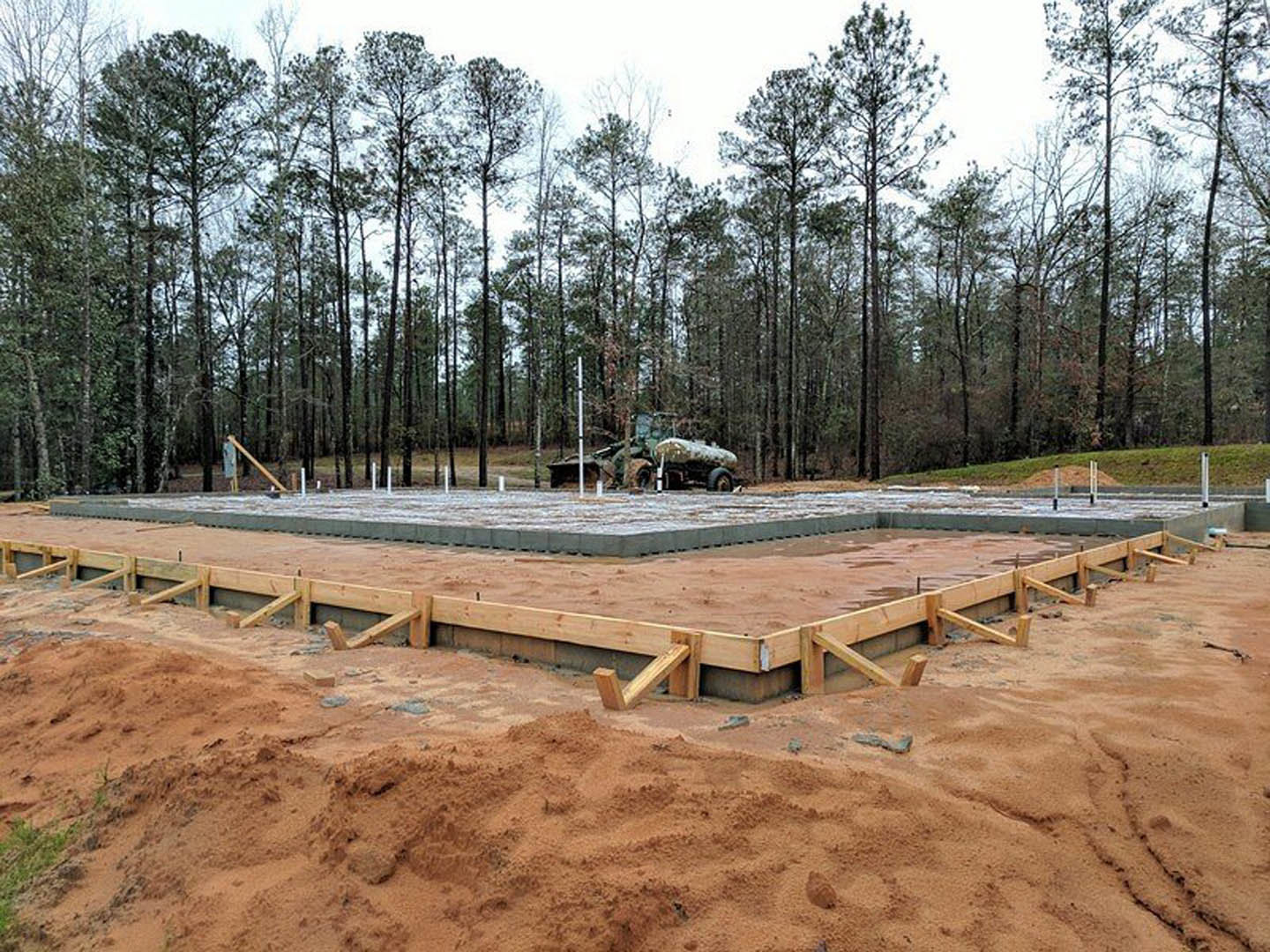Wooden foundation and beams on a construction site surrounded by trees, with a truck parked in the background and a pile of sand nearby.