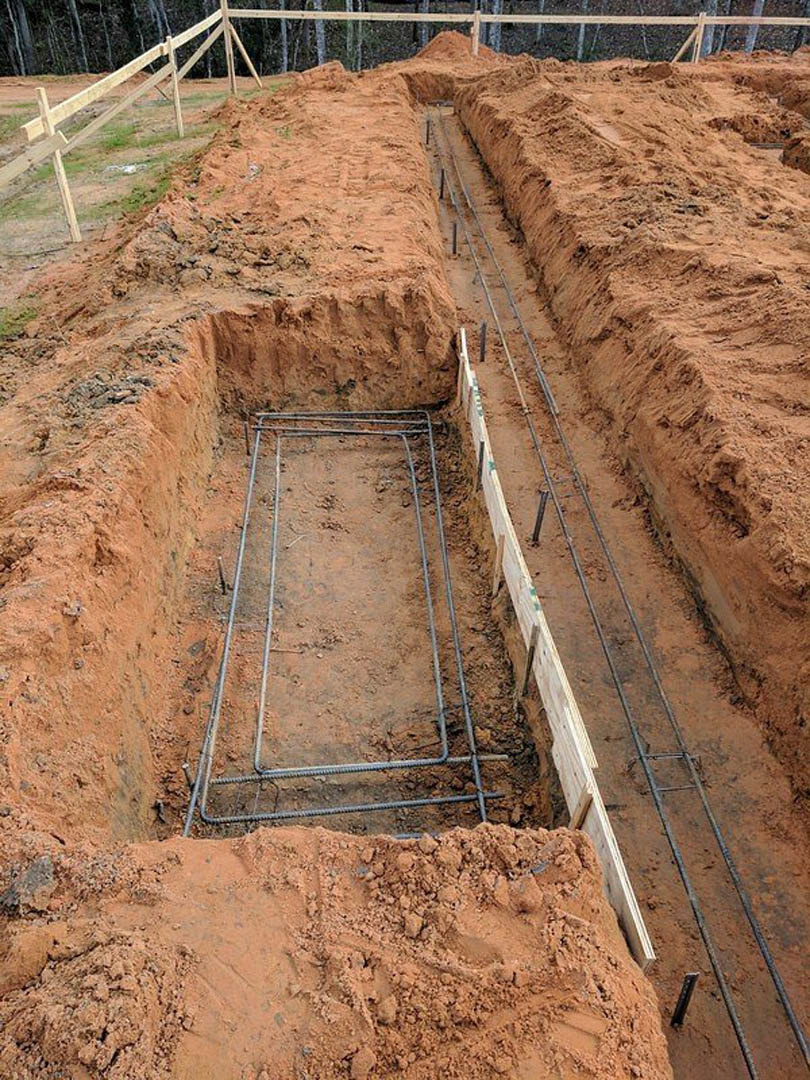 Trench filled with metal rods and soil at a residential construction site, bordered by a wooden fence and piles of dirt