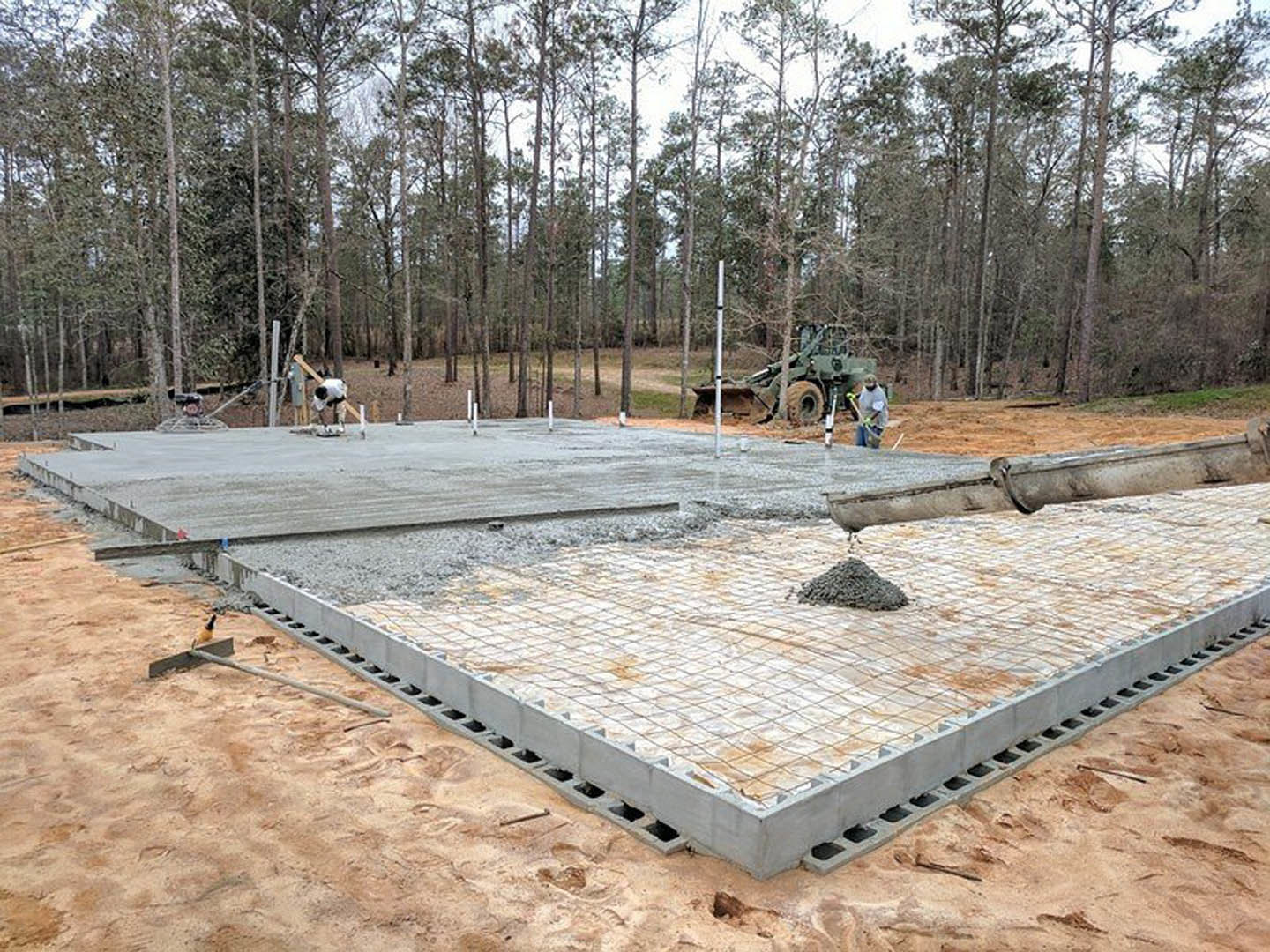 Concrete foundation slab surrounded by soil and trees, man in work clothes standing beside partially built custom home structure
