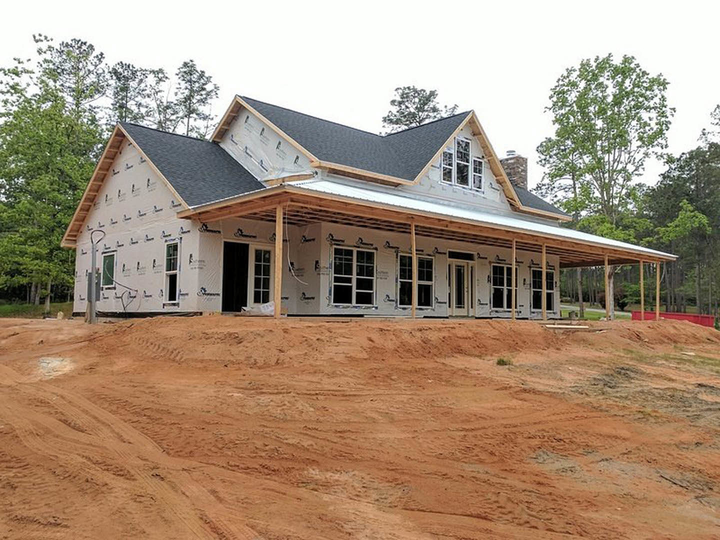 Partially built house with exposed roof framing, surrounded by dirt and mature trees, Karen Blixen Museum visible in the background