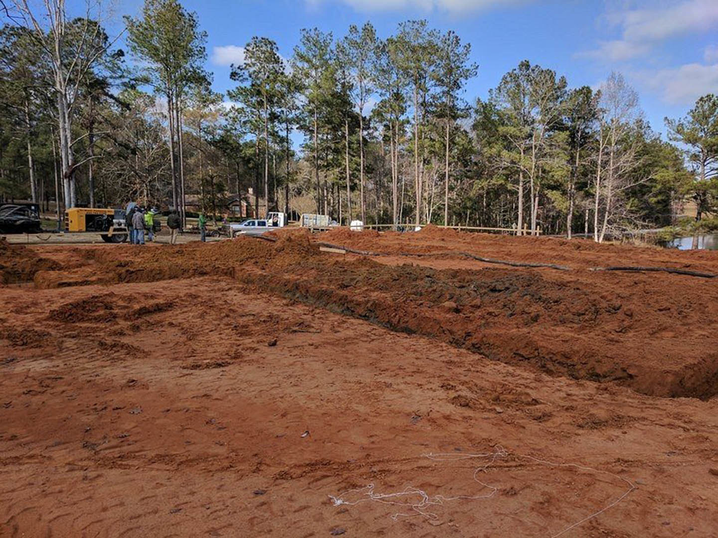 Dirt field bordered by leafy trees, scattered people, yellow truck parked near a pile of soil, cloudy sky overhead