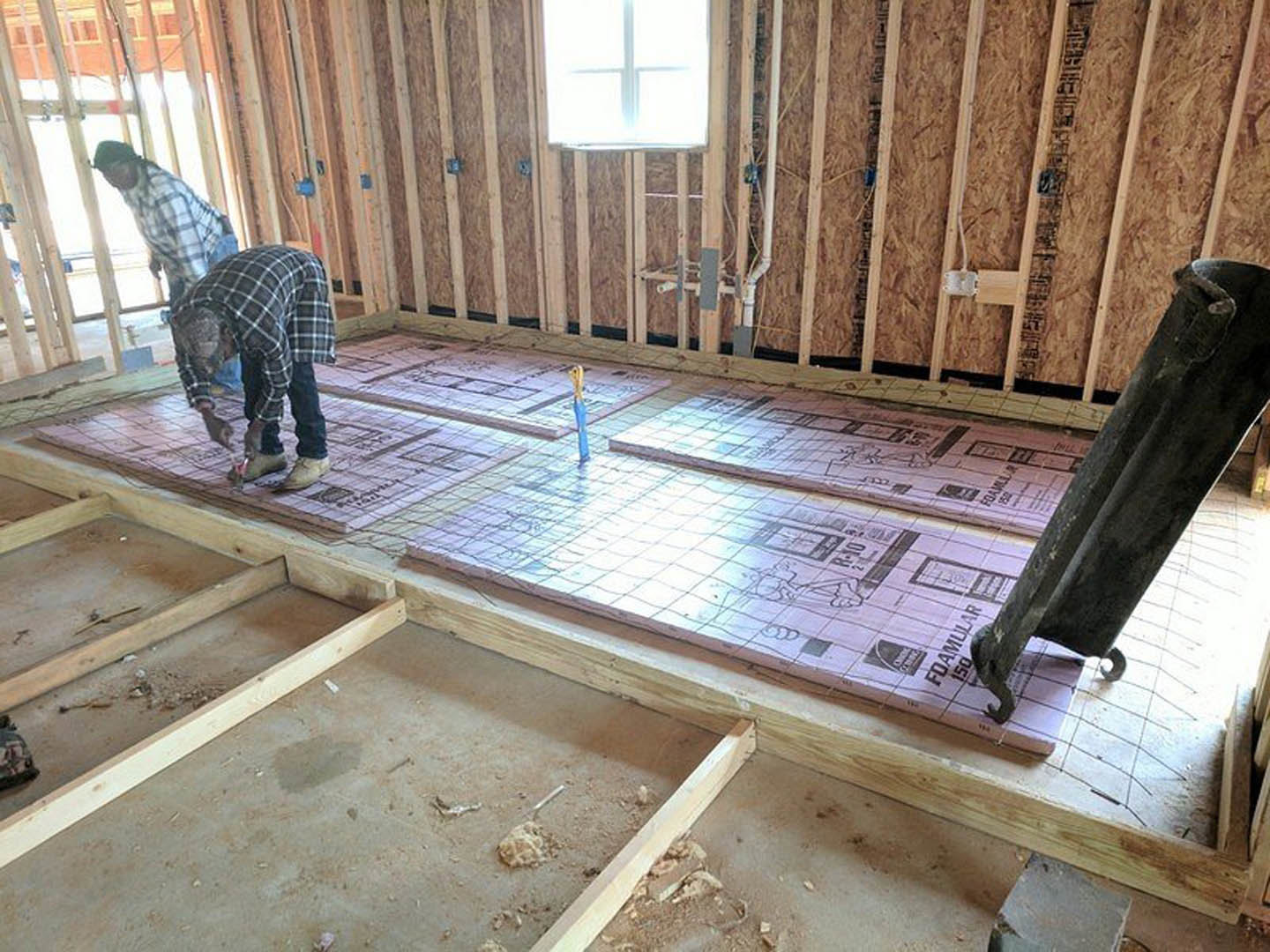 Two men installing flooring in a room with a wooden post, tile floor, and sunlight streaming through a window