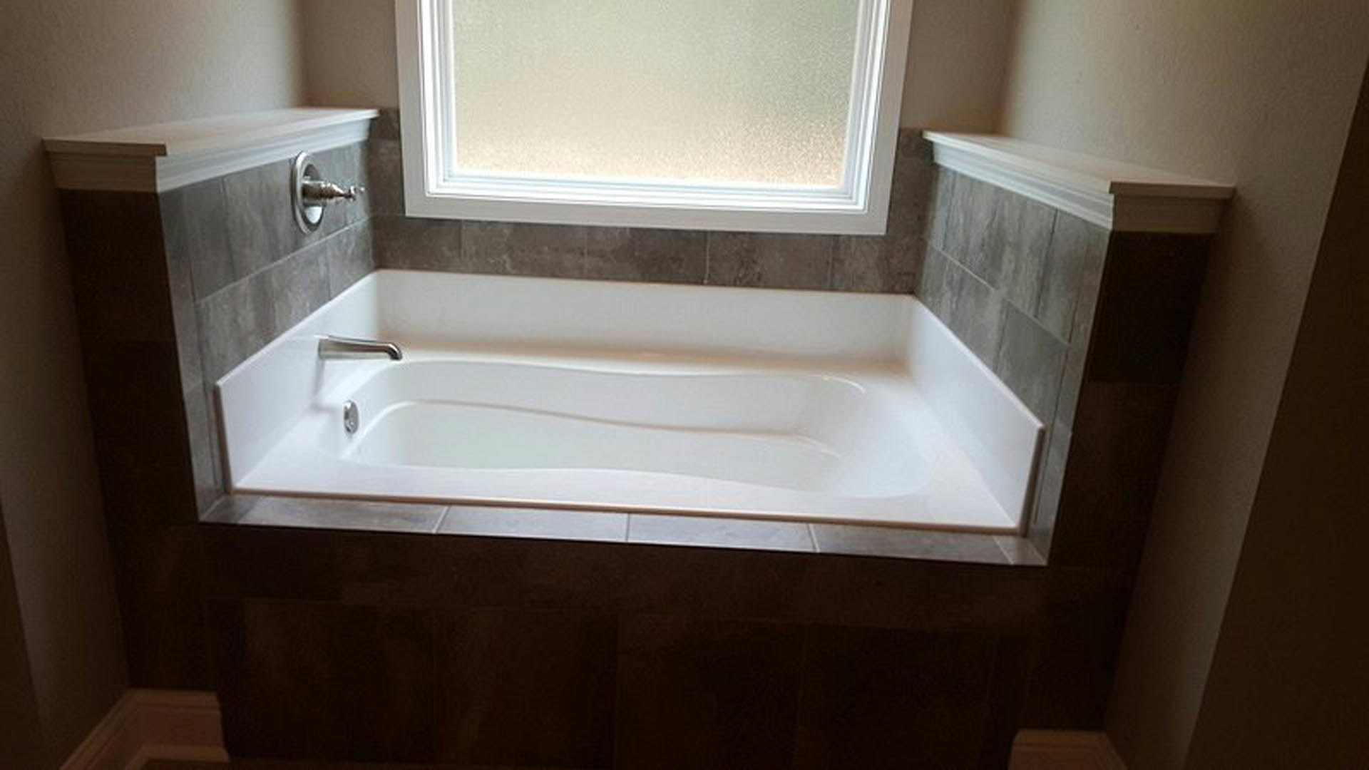White freestanding bathtub with chrome faucet beside large window, surrounded by light gray tile walls and floor