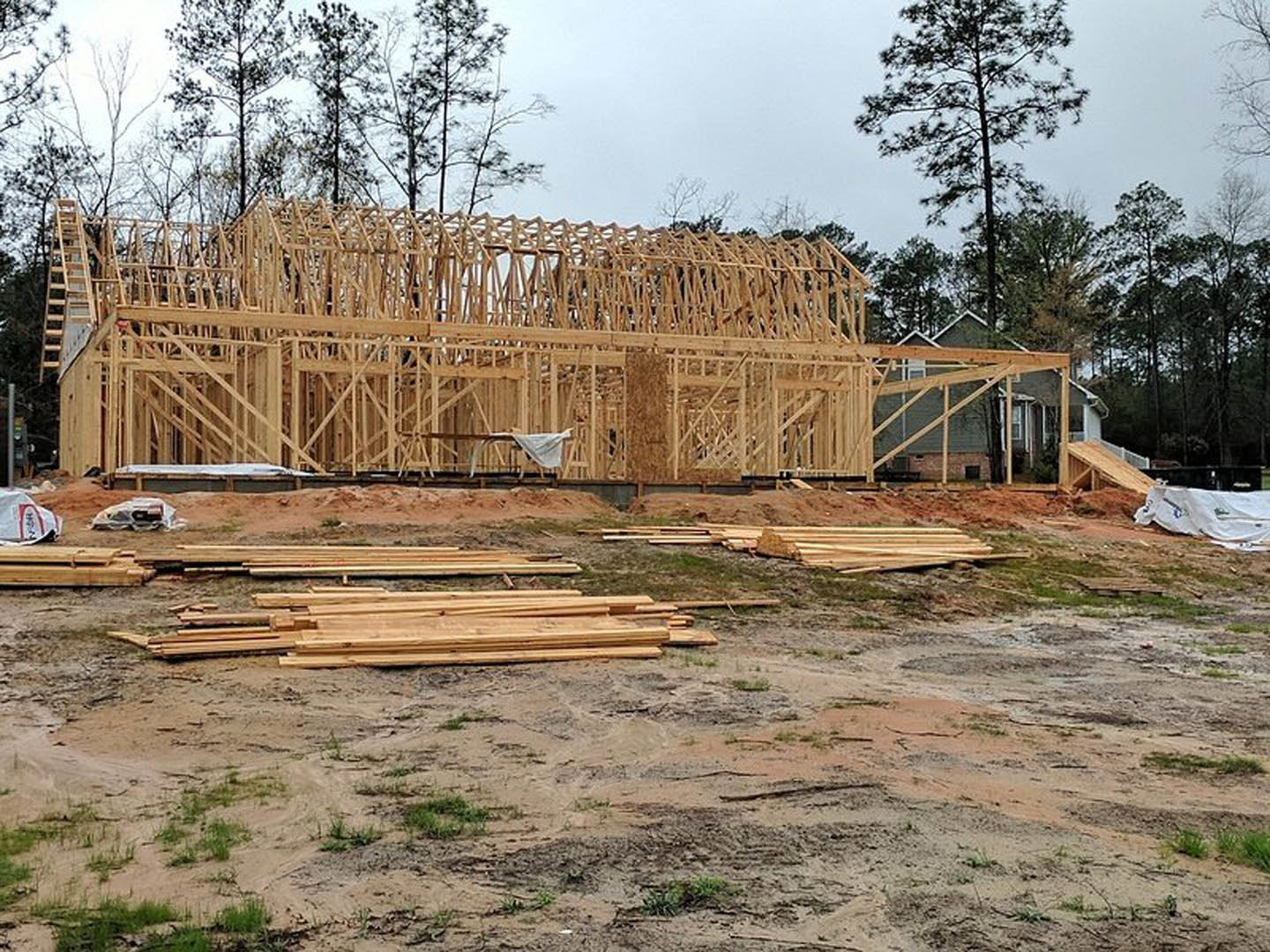 Framed wooden house under construction with exposed beams, piles of lumber on the ground, tall tree nearby, and partially installed roof with white railing.