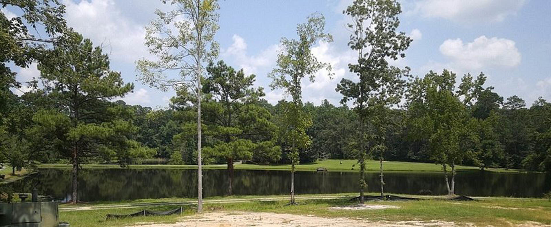 Modern lakeside home with large windows, surrounded by green grass and mature trees, overlooking calm water and blue sky with scattered clouds