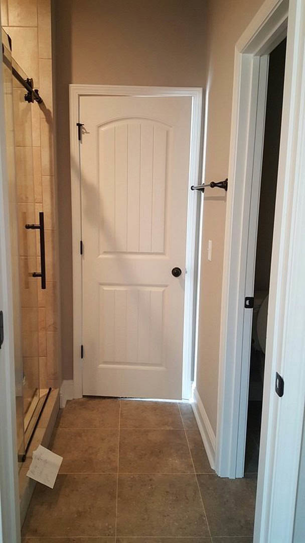 White paneled bathroom door with black knobs, brown tile floor with white grout, partial view of wall