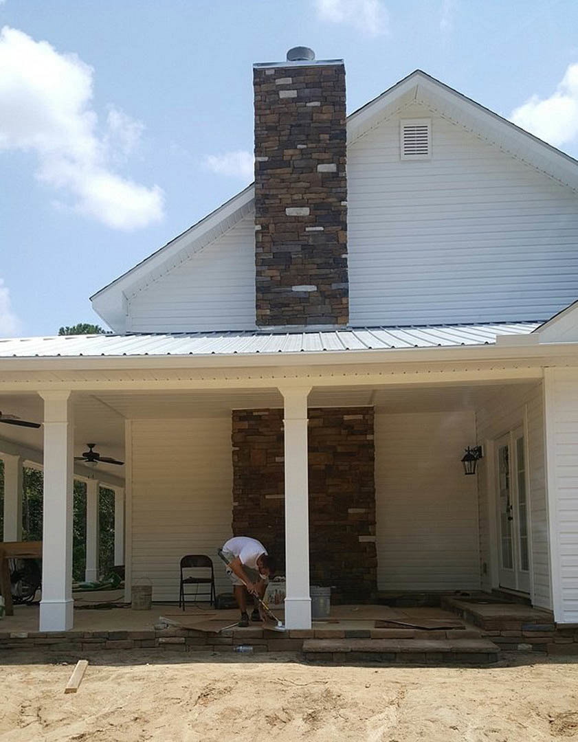 Man using a tool on covered porch with black chair, wooden seat, brick chimney, white siding, and white roof vent