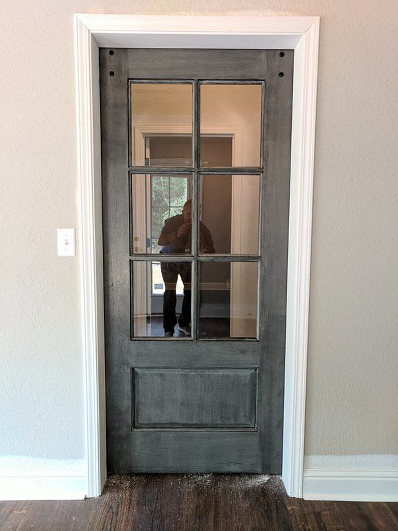 Man standing in a doorway with wood door and metal handle, light streaming through nearby window onto hardwood floor and neutral walls