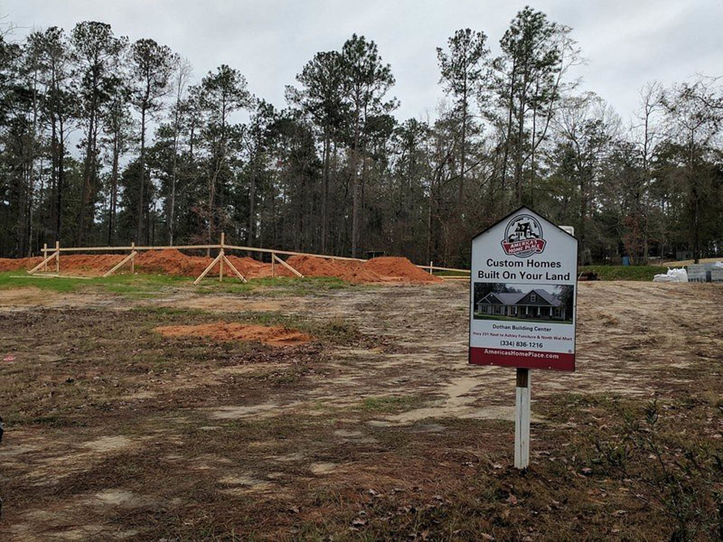 Wooden post with a house-shaped sign beside a dirt lot, bordered by grassy field and dense trees under a partly cloudy sky