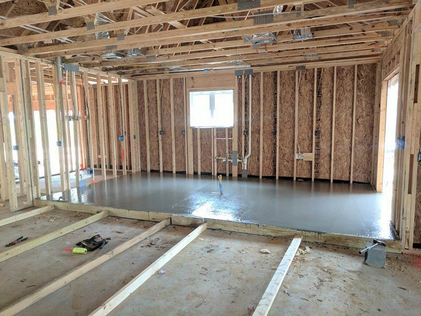Concrete floor with sunlight streaming through a large window, exposed wooden ceiling beams, construction tools and materials scattered on unfinished surfaces.