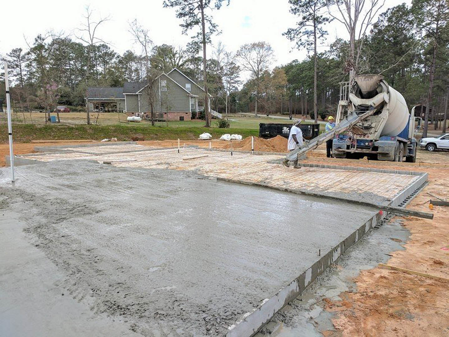 Concrete mixer truck pouring fresh concrete onto a slab foundation at a residential construction site, with a worker guiding the flow and trees visible in the background.
