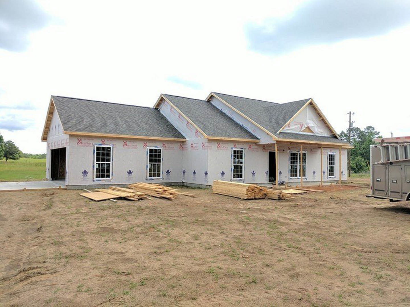 Wood-framed house under construction with exposed beams and roof, grassy yard, multi-pane windows, Robert Frost Farm visible in background under cloudy sky