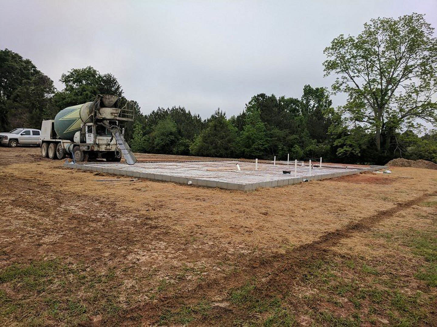 Concrete mixer truck with green and yellow drum parked beside newly poured concrete foundation slab, surrounded by grassy field and leafy trees under partly cloudy sky