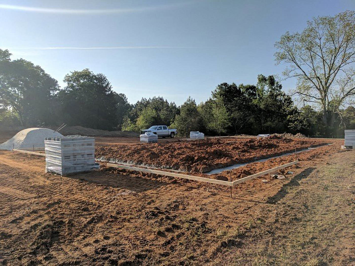 Dirt construction site bordered by trees, wooden beam lying on soil, pallet wrapped in plastic, large white structure with fence in background, white car parked along road, cloudy