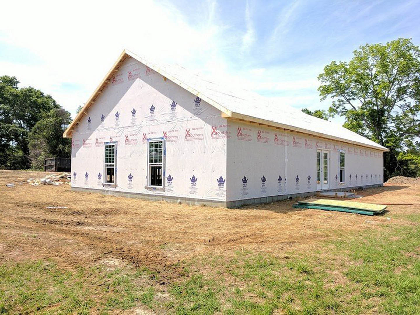 Two-story house under construction with exposed wooden framing, partially finished roof, and patch of bare dirt in front yard