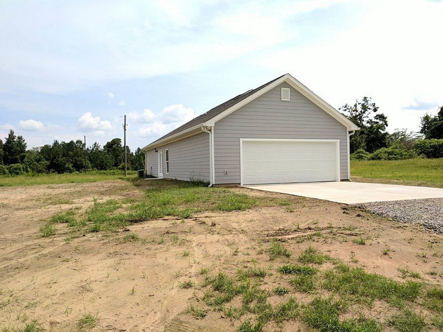 Grey garage with white paneled door, concrete driveway, dirt patch, white pole, and grey shingle roof; grassy lot with trees and cloudy sky in background.